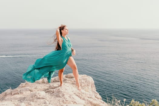 Side view a Young beautiful sensual woman in a mint long dress posing on a volcanic rock high above the sea during sunset. Girl on the nature on overcast sky background. Fashion photo