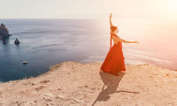 Side view a Young beautiful sensual woman in a red long dress posing on a rock high above the sea during sunrise. Girl on the nature on blue sky background. Fashion photo.