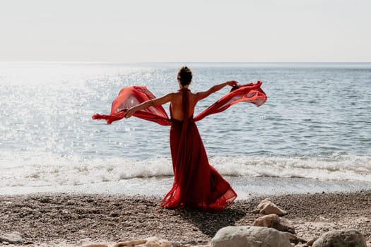 Side view a Young beautiful sensual woman in a red long dress posing on a rock high above the sea during sunrise. Girl on the nature on blue sky background. Fashion photo.