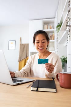 Happy young Asian woman online shopping using laptop and credit card at home office. Vertical image. E-commerce concept.