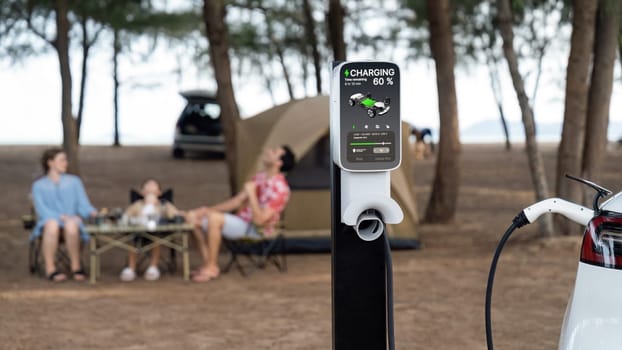 Alternative family camping trip traveling by the beach with electric car recharging battery from EV charging station with blurred family enjoying the seascape campsite background. Perpetual