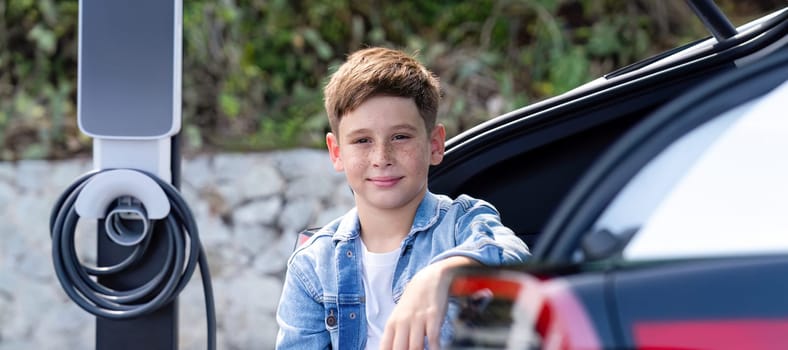 Little boy sitting on car trunk while recharging eco-friendly electric car from EV charging station. EV car road trip travel with alternative vehicle powered sustainable energy. Panorama Perpetual