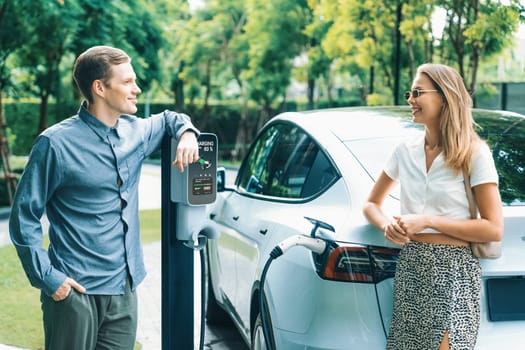 Young couple travel with EV electric car charging in green sustainable city outdoor garden in summer shows urban sustainability lifestyle by green clean rechargeable energy of electric vehicle innards