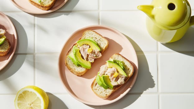 Slices of bread topped with tuna and avocado are arranged on a pale plate. A bright yellow teapot and lemon complement the midday setting and add warmth to the atmosphere.