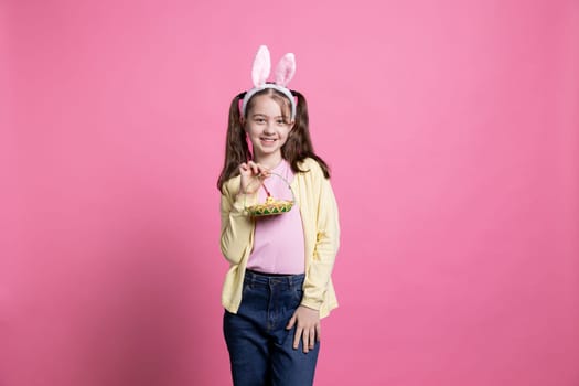 Charming little girl holding basket of eggs and other easter toys, pink background. Toddler expressing childhood innocence and the joyous anticipation of spring holiday festivities.