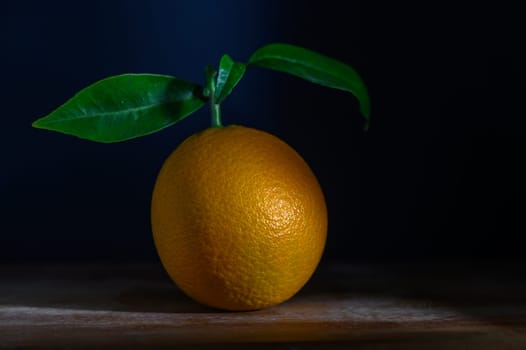 A bright orange sits elegantly on a wooden surface, its green leaf adding a touch of vitality against a dark background.