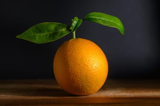 A luscious orange rests on a wooden surface, displaying its rich color and fresh green leaves in warm light.
