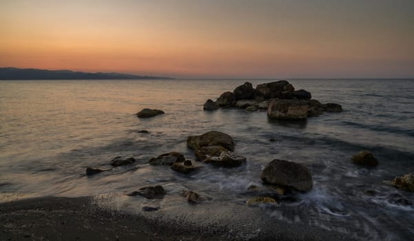 Waves gently lap against the rocky shore under a peaceful twilight sky, reflecting soft colors of sunset.