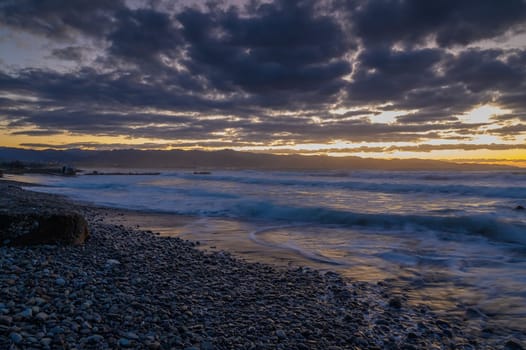 Golden hues reflect on water while waves gently roll over pebbles under a dramatic sky at sunset