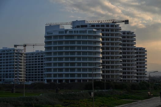 Newly built apartment structures rise amidst construction cranes as the sun sets, casting shadows over the landscape.