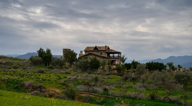 Nestled in the hills, a rustic villa stands amidst vibrant green fields and distant mountains under a dramatic sky.