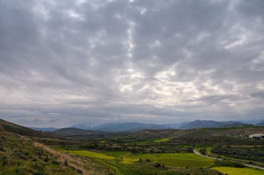 Lush green fields and winding roads contrast with overcast skies and distant peaks in a tranquil landscape.
