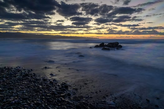 Waves softly lap at smooth stones along the shore as the sun sets, painting the sky with vibrant colors and clouds.