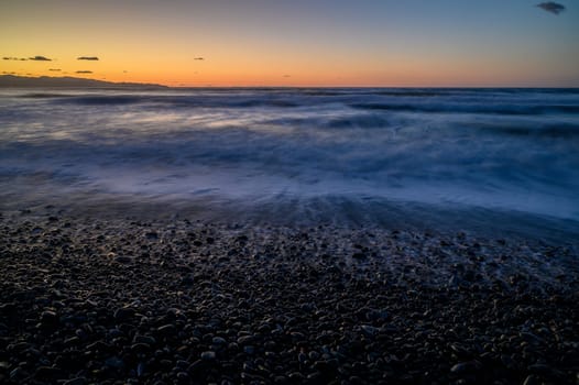 Gentle waves roll over smooth pebbles under a vivid twilight sky, creating a peaceful coastal atmosphere at sunset.