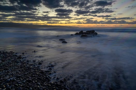 Waves kiss the rocky shore as twilight turns the sky into tranquil shades of orange and blue.