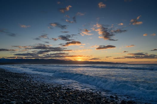 Golden sunlight reflects on the water while waves peacefully roll onto a pebble-strewn coastline at dusk.