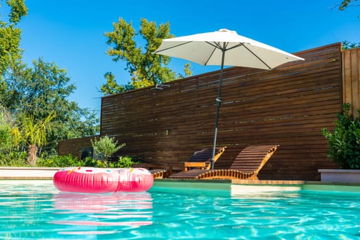 Turquoise backyard swimming pool with pink inflatable ring, wooden deck, umbrella, and lounge chairs