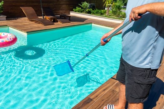 Man using skimmer net to clean turquoise swimming pool water with wooden deck and colorful floaties