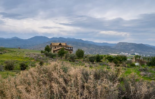 A charming stone house quietly rests amidst lush hills, framed by distant mountains and a dramatic sky at twilight.