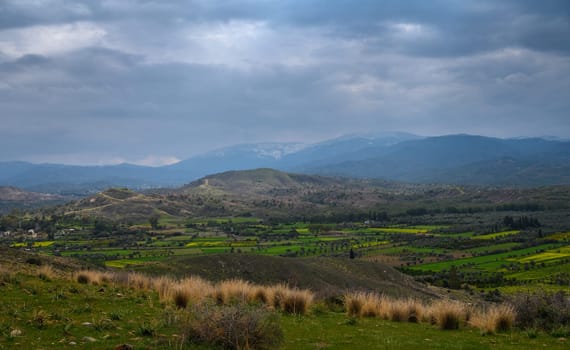 Beneath a moody sky, verdant hills stretch across a tranquil valley filled with vibrant fields and distant mountains.