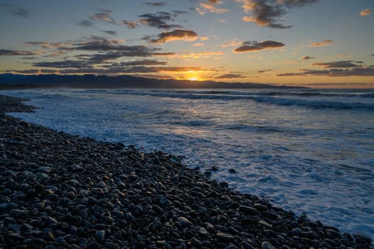 Golden sun descends on the horizon, illuminating pebbled beach while waves embrace the shore at twilight.