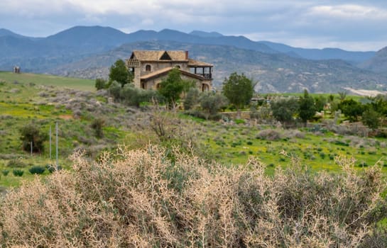 A rustic house blends seamlessly into a vibrant green landscape, surrounded by mountains under a cloudy sky.