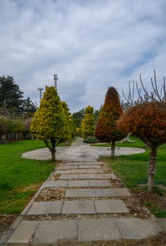A well-maintained pathway winds through lush greenery, framed by colorful, carefully shaped trees under a cloudy sky.