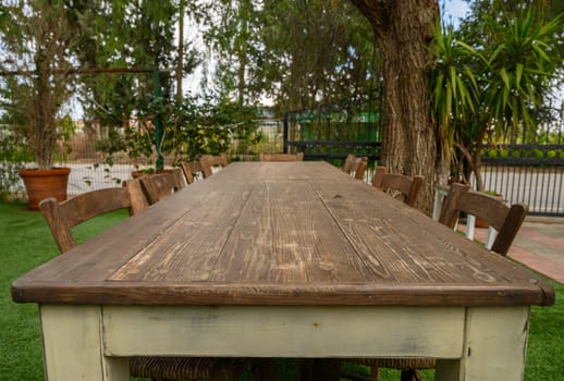 A weathered wooden table lined with rustic chairs sits in a lush garden bathed in warm sunlight.