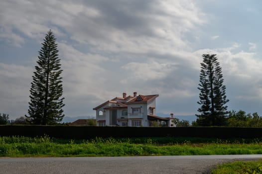 A beautiful villa stands amidst lush greenery, flanked by towering trees, as clouds drift across the blue sky.