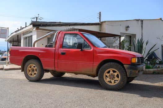 A vibrant red pickup truck rests on a gravel road beside a simple, weathered structure in the sunlight.