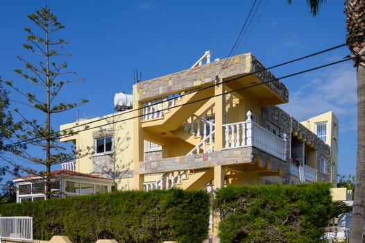 A colorful Mediterranean villa stands against a bright blue sky, surrounded by flourishing plants and palm trees.