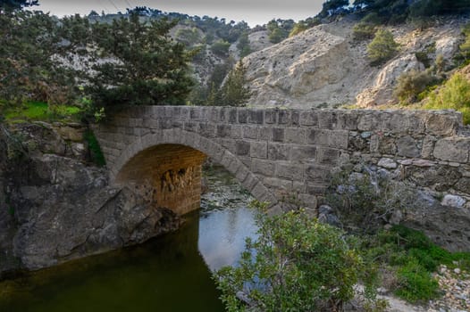 A picturesque stone bridge arches gracefully over crystal-clear water, surrounded by scenic hills and greenery.