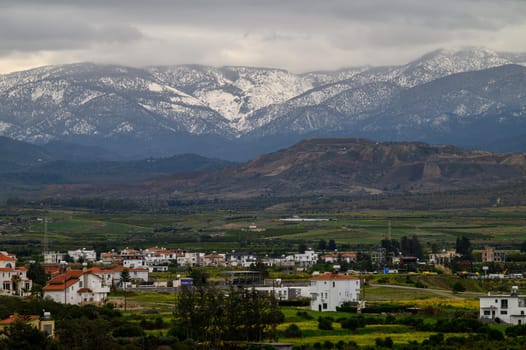 Snow-capped peaks tower over a quaint village, with lush greenery and fields stretching towards the horizon.