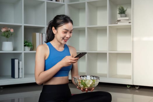 A woman enjoying a colorful salad and using her smartphone while sitting in a stylish living area, blending healthy eating with technology.