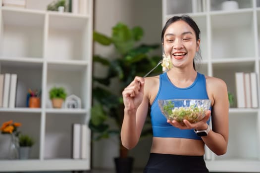 A cheerful woman in athletic clothing happily prepares to eat a nutritious salad, promoting healthy living in a stylish home environment.