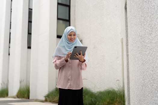 A smiling Muslim businesswoman engages with her tablet outdoors, showcasing a blend of modern work culture and cultural diversity in a contemporary environment.