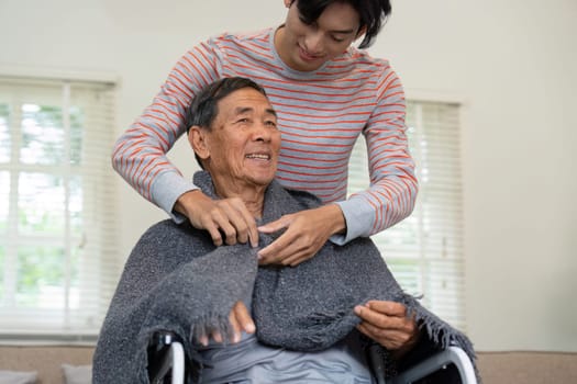 A son assists his elderly father in adjusting a blanket while he sits in a wheelchair, emphasizing care and family support in a comforting home atmosphere.