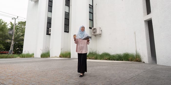 A confident Muslim businesswoman walks through an urban setting, holding a tablet and coffee, symbolizing modern professionalism and cultural diversity.