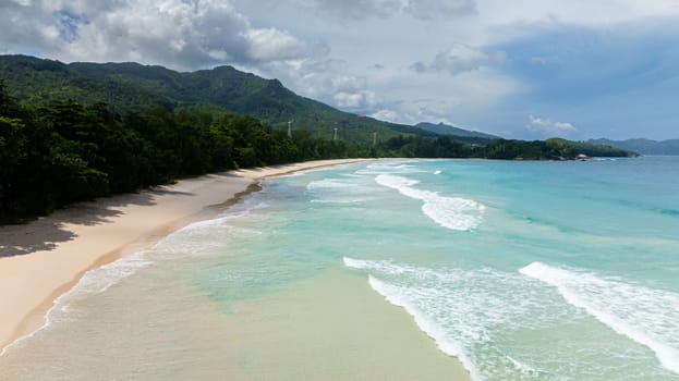 White sandy beach meets turquoise waves, surrounded by tropical vegetation and greenery. Seychelles, Mahe. Grand Anse Beach.