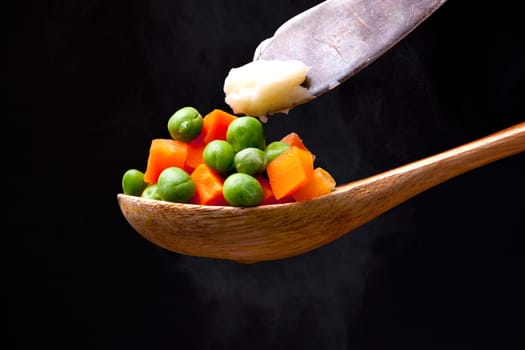 Peas and carrots topped with butter on a wooden spoon with steam from below.