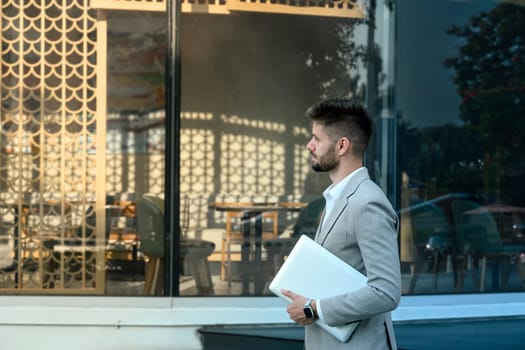 Businessman holding laptop walking outside modern office building.