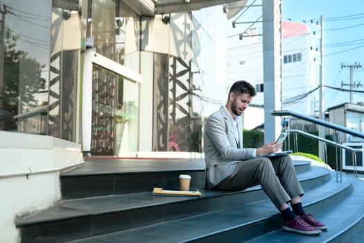 Young professional in suit checking smartphone sitting outdoors at office building.