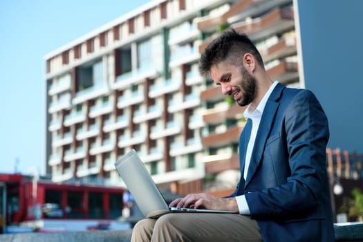 Smiling bearded businessman in formal suit working on laptop outdoors.