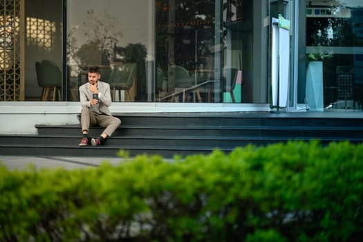 Young professional in suit checking smartphone sitting outdoors at office building.