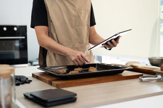 Small business owner checking orders on digital tablet while preparing cookie dough in modern kitchen.