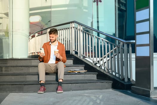 Young businessman sitting on outdoor office stairs and checking mobile phone.
