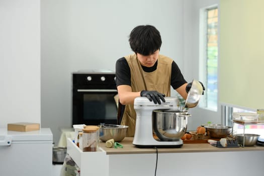 Man wearing black gloves pouring flour into stand mixer while preparing cookie dough.