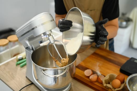 Man wearing black gloves pouring flour into stand mixer while preparing cookie dough.