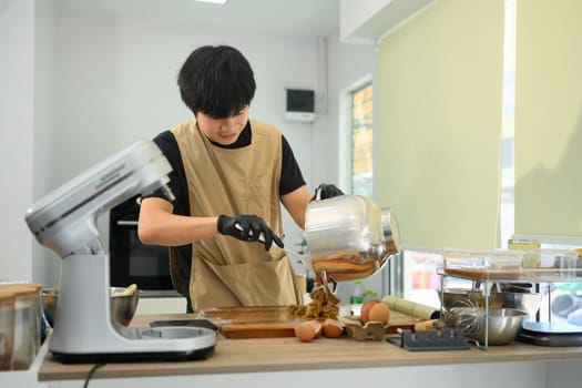 Young man adding ingredients into a stand mixer while preparing cookie dough.