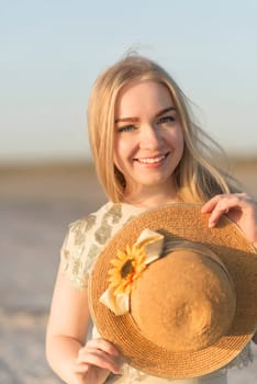 Close-up of a blonde girl with a straw hat in her hands, she laughs and looks at the camera. The hat is decorated with sunflowers. Summer, sunny weather